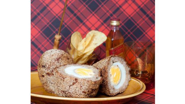 Ein aufgeschnittenes Scotch Egg liegt auf einem Teller, zusammen mit Chips und einer Flasche Whisky.