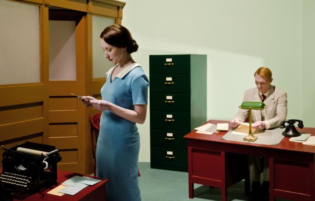 In einem Büro liest eine Frau in blauem Kleid einen Brief, während ein Mann am Schreibtisch sitzt.