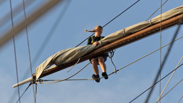 Eine junge Frau sitzt auf einem Mast eines Segelschiffs.