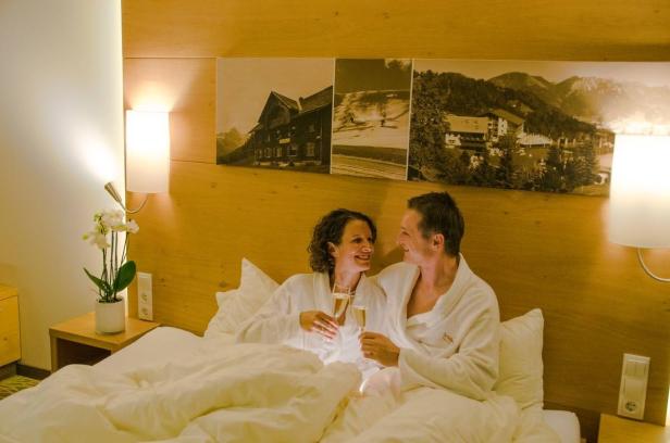 Ein Paar in Bademänteln liegt im Bett und prostet sich mit Sekt zu.