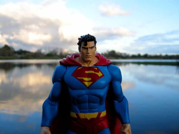 Eine Superman-Actionfigur steht vor einem See unter bewölktem Himmel.
