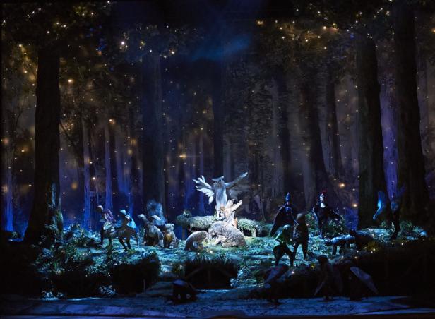 Eine Theaterszene mit Darstellern und Tierfiguren in einem dunklen Wald.