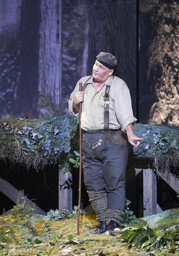 Ein Mann in Tracht mit Wanderstock steht vor einer Waldkulisse.