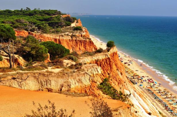 Blick auf einen Strand mit roten Klippen und vielen Sonnenschirmen.