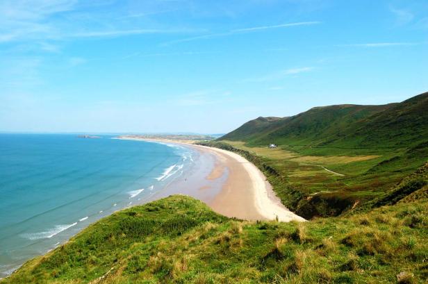 Blick von einem grünen Hügel auf einen langen Sandstrand und das Meer.