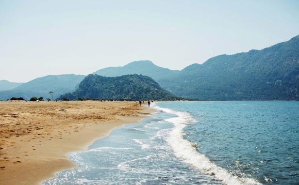 Ein Sandstrand mit blauem Wasser und bewaldeten Bergen im Hintergrund.
