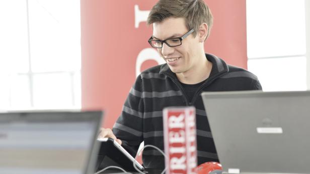 Ein Mann mit Brille lächelt, während er ein Tablet benutzt; Laptops sind im Vordergrund zu sehen.