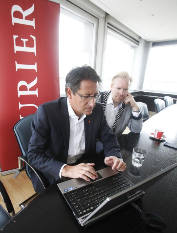 Zwei Männer sitzen an einem Tisch, einer arbeitet am Laptop, im Hintergrund ein „Kurier“-Banner.