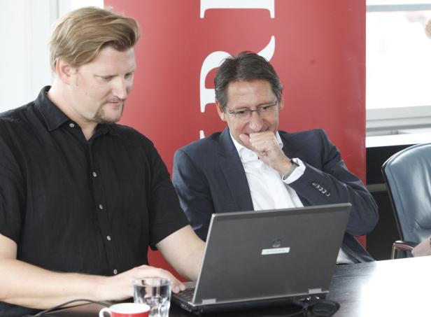Zwei Männer sitzen an einem Tisch und schauen auf einen Laptop.
