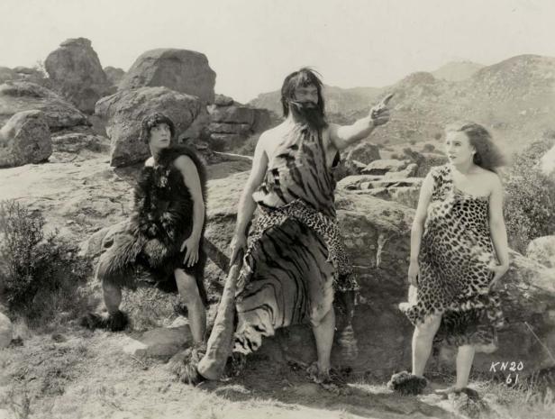 Drei in Tierfelle gekleidete Personen stehen in einer felsigen Landschaft.