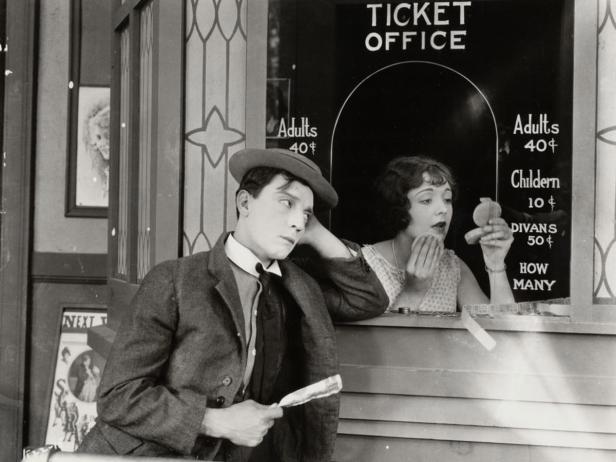 Ein Mann wartet gelangweilt vor dem Ticketschalter, während eine Frau sich schminkt.