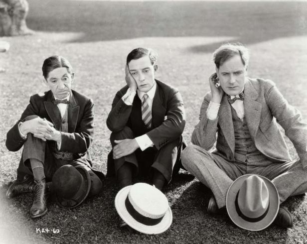 Drei Männer in Anzügen sitzen mit Hüten auf dem Rasen.