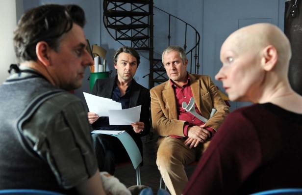 Vier Personen sitzen in einem Raum, zwei Männer halten Papiere, eine Frau hat eine Gehhilfe.