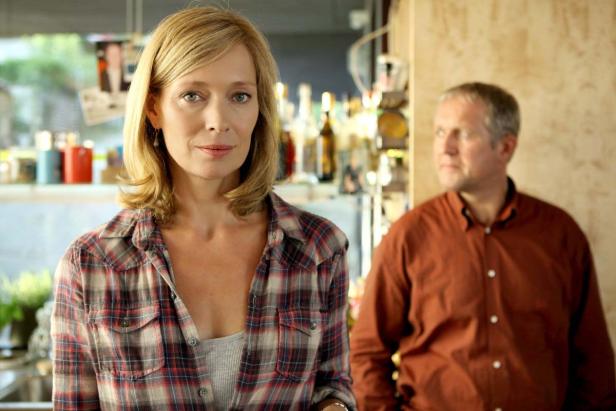 Eine Frau mit kariertem Hemd und ein Mann im Hintergrund stehen in einem Raum.