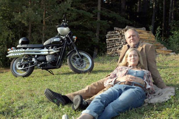 Ein Paar entspannt auf einer Wiese neben einem Motorrad und einem Holzstapel.