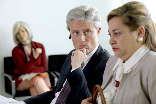 Ein Mann in einem Anzug sitzt mit zwei Frauen in einem Wartezimmer.