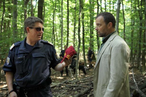 Ein Polizist hält eine rote Kappe mit der Zahl 93 und spricht mit einem Mann im Wald.