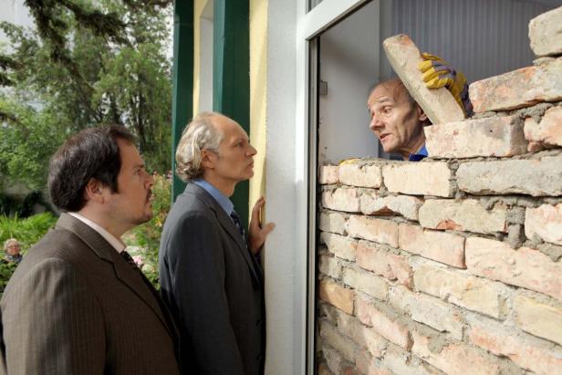 Zwei Männer in Anzügen beobachten einen Maurer, der eine Wand hochzieht.