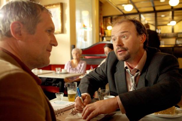 Zwei Männer sitzen in einem Café, einer zeichnet in ein Notizbuch.