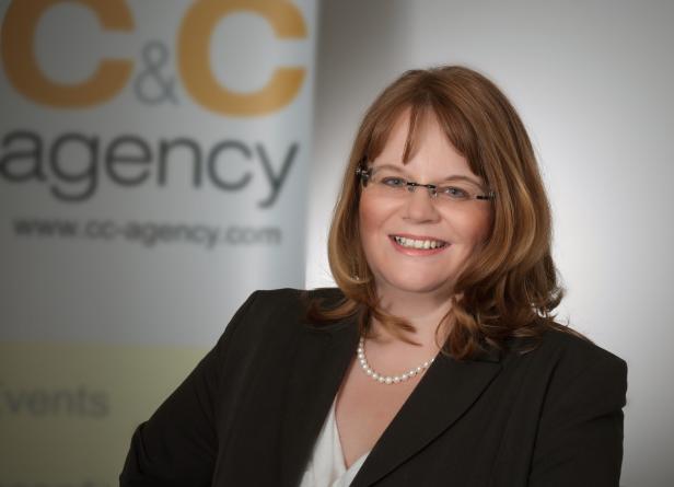 Eine lächelnde Frau mit Brille und dunklem Blazer vor dem Logo der C&C Agency.