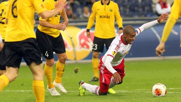 Ein Fußballspieler in rot-weißer Spielkleidung rutscht auf dem Rasen, während andere Spieler in gelben Trikots zusehen.