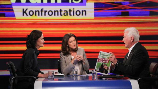 Drei Personen sitzen an einem Tisch vor dem Hintergrund einer Fernsehsendung zum Thema „Konfrontation“.