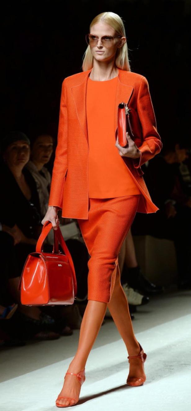 Eine Frau in einem orangefarbenen Hosenanzug mit passender Handtasche auf dem Laufsteg.