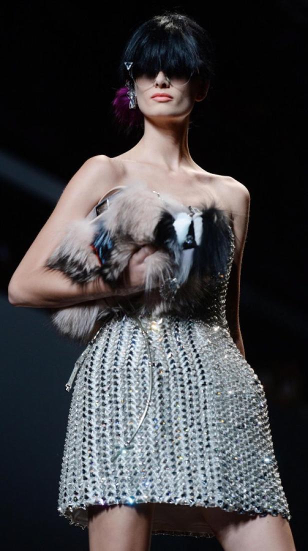 Ein Model mit Ponyfrisur und Brille präsentiert ein silbernes Kleid und eine Fellhandtasche.