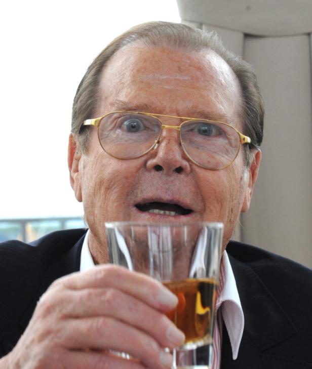 Roger Moore hält ein Glas mit einem alkoholischen Getränk in der Hand.