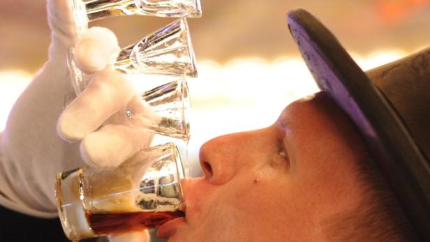 Ein Mann mit Hut trinkt aus einer Reihe von Schnapsgläsern, die von einer Hand mit Handschuh gehalten werden.