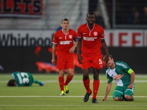 Ein Fußballspieler in Rot steht auf dem Feld, während zwei andere Spieler am Boden liegen.