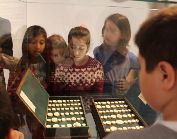 Kinder betrachten in einem Museum zwei offene Kästen mit alten Münzen.