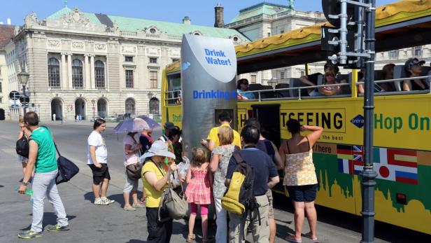 Hitze Touristen stehen an einem Trinkwasserbrunnen neben einem gelben Hop-On-Bus in Wien.