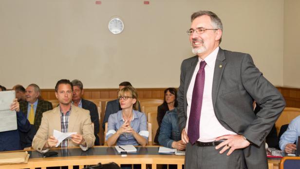 Ein Mann im Anzug steht vor einer Gruppe von Menschen in einem Gerichtssaal oder Sitzungssaal.