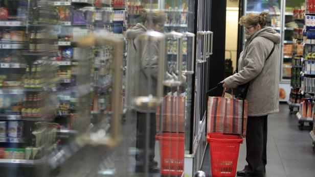 Eine Frau steht mit Einkaufstasche und -korb vor einem Kühlregal in einem Supermarkt.