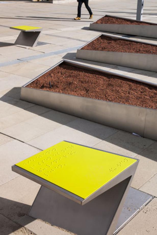 Eine gelbe Bank mit Braille-Schrift steht auf einem Platz, im Hintergrund sind bepflanzte Hochbeete zu sehen.