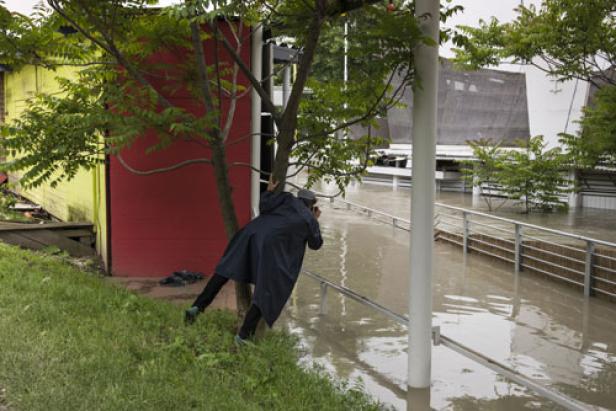 Eine Person in Regenkleidung fotografiert das Hochwasser, das Gebäude und Zäune überflutet hat.