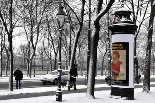 Winterliche Straßenszene in Wien mit einer Litfaßsäule und Passanten.