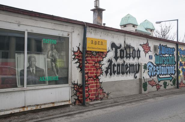 Die Fassade eines Gebäudes mit Graffiti, darunter „Tattoo Academy“ und „Wiener Würschtl Restaurant“.