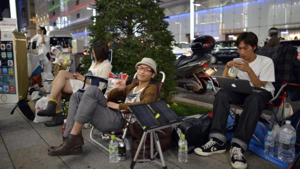 Menschen sitzen auf Stühlen vor einem Geschäft in Tokio und nutzen ihre elektronischen Geräte.