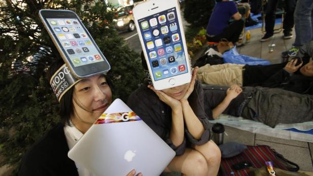 Zwei Personen mit iPhone-Attrappen auf dem Kopf und einem MacBook in der Hand sitzen vor einem Geschäft.