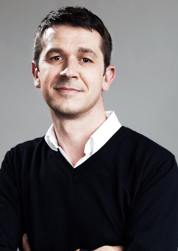 Ein Mann mit dunklen Haaren und einem schwarzen Pullover vor grauem Hintergrund.