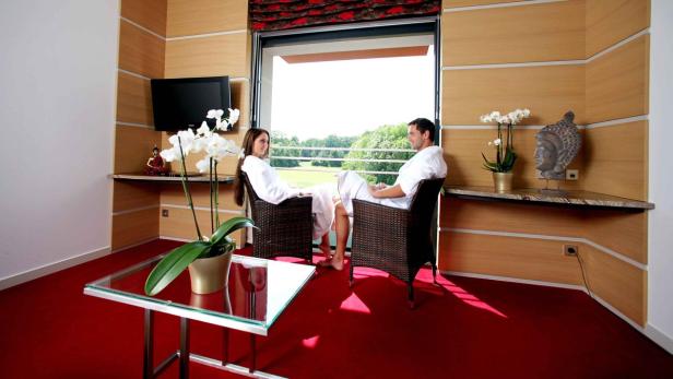 Ein Paar in Bademänteln sitzt in einem Hotelzimmer mit Blick auf eine grüne Landschaft.