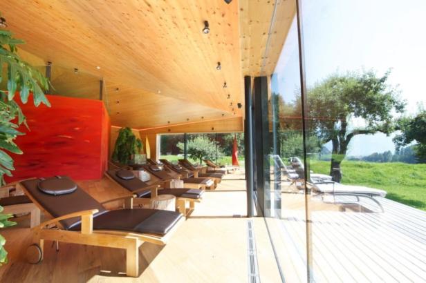 Ein Innenraum mit Liegestühlen und Blick durch eine Glaswand auf eine grüne Wiese.