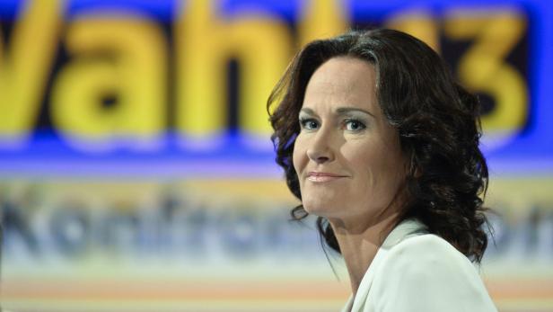 Eine Frau mit dunklen Haaren vor einem unscharfen Hintergrund mit der Aufschrift „Wahl 13“.