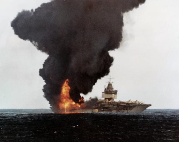Ein Flugzeugträger steht in Flammen auf dem Meer, dichte Rauchwolken steigen auf.