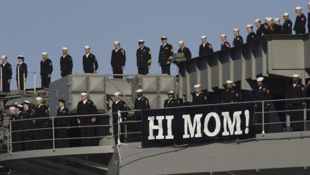 Marineangehörige stehen an Deck eines Schiffes mit einem Banner „Hi Mom!“.
