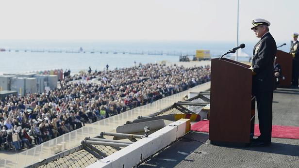 Ein Marineoffizier hält eine Rede vor einer großen Menschenmenge am Meer.