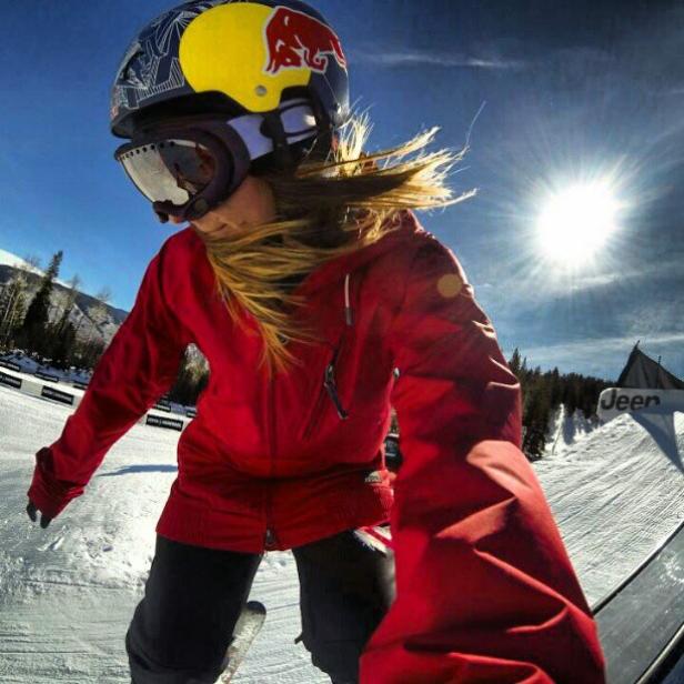 Eine Snowboarderin mit roter Jacke und Red Bull Helm fährt auf einer schneebedeckten Piste.