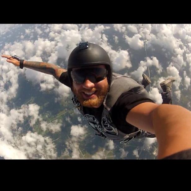 Ein lächelnder Mann mit Helm und Sonnenbrille springt Fallschirm über den Wolken.